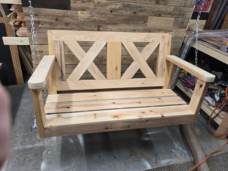 May include: A wooden swing bench with a slatted seat and a criss-cross design on the back. The swing is made of unfinished wood and is hanging from chains.