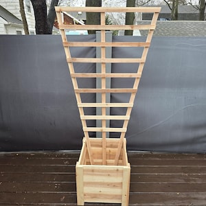 May include: A wooden planter box with an attached trellis. The planter box is rectangular and made of light-coloured wood. The trellis is a tall, open structure, also made of wood, with a lattice design. The background includes a grey privacy screen.
