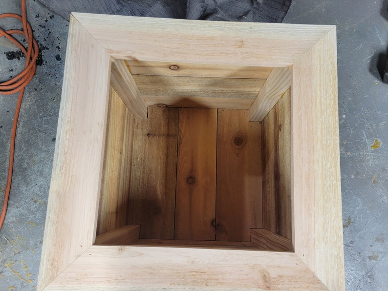 K&ouml;nnte beinhalten: Ein quadratischer Pflanzenkasten aus Holz mit einer nat&uuml;rlichen Holzoptik. Der Pflanzenkasten ist leer und bereit, mit Pflanzen bef&uuml;llt zu werden.