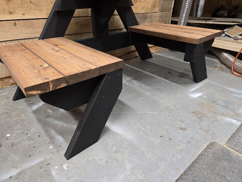 May include: A black wooden picnic table with two benches. The table and benches are made of wood with a dark brown stain. The table is in a workshop setting.