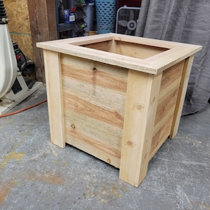 K&ouml;nnte beinhalten: Ein quadratischer Holz-Pflanzkasten aus hellem braunem Holz mit nat&uuml;rlicher Oberfl&auml;che. Der Pflanzkasten hat ein schlichtes Design mit einem erh&ouml;hten Rand an der Oberseite.
