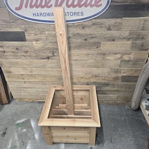 May include: A wooden planter box with a square shape and a raised edge. The planter is made of unfinished wood and has a natural wood color. A wooden post is standing upright in the center of the planter.