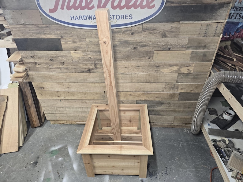 May include: A wooden planter box with a square shape and a raised edge. The planter is made of unfinished wood and has a natural wood color. A wooden post is standing upright in the center of the planter.