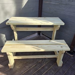 May include: A wooden bench and table set made of unfinished wood. The table has a simple design with a single shelf underneath. The bench is also simple with a single seat.