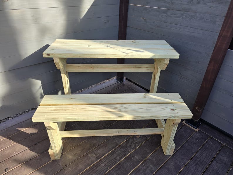 May include: A wooden bench and table set made of unfinished wood. The table has a simple design with a single shelf underneath. The bench is also simple with a single seat.