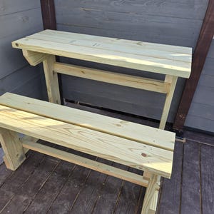 May include: A wooden bench and table set made of light brown wood. The bench is attached to the table and has a simple design. The table is a bit taller than the bench.