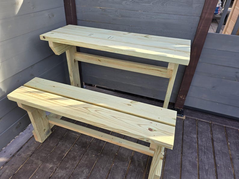May include: A wooden bench and table set made of light brown wood. The bench is attached to the table and has a simple design. The table is a bit taller than the bench.
