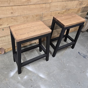 Puede incluir: Dos taburetes de metal negro con tapas de madera. Los taburetes tienen un dise&ntilde;o sencillo con un asiento rectangular y un solo estante debajo.