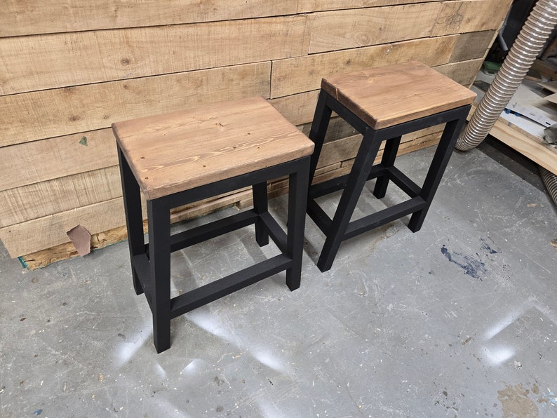 Puede incluir: Dos taburetes de metal negro con tapas de madera. Los taburetes tienen un dise&ntilde;o sencillo con un asiento rectangular y un solo estante debajo.