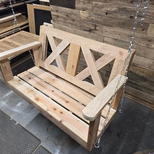 May include: A wooden swing bench with a slatted seat and a decorative X-shaped back. The swing is hanging from two silver chains.