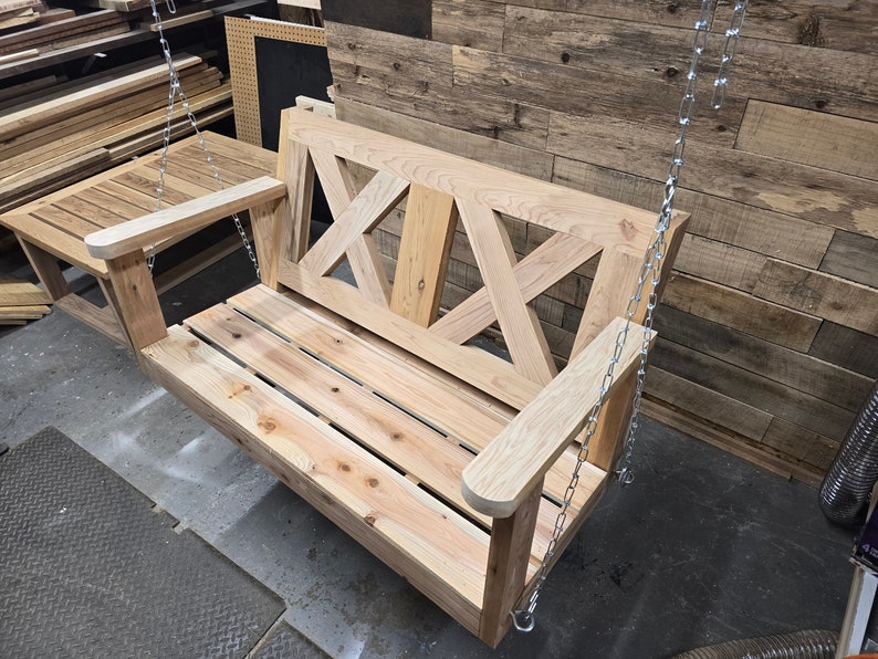 May include: A wooden swing bench with a slatted seat and a decorative X-shaped back. The swing is hanging from two silver chains.