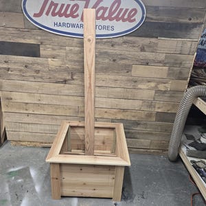 May include: A wooden planter box with a tall, vertical wooden post in the center. The planter box is made of unfinished wood and has a square shape. The post is also made of unfinished wood and is taller than the planter box. The planter box is sitting on a wooden floor.