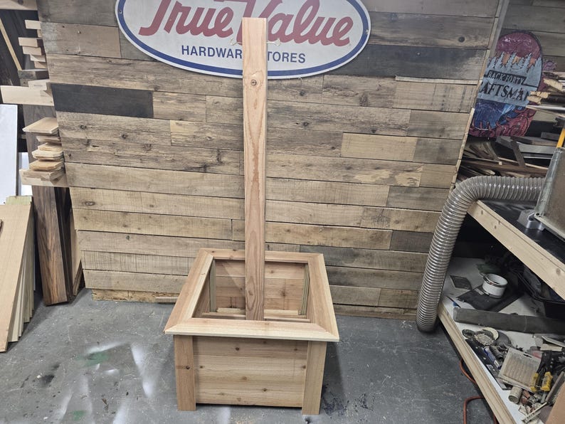 May include: A wooden planter box with a tall, vertical wooden post in the center. The planter box is made of unfinished wood and has a square shape. The post is also made of unfinished wood and is taller than the planter box. The planter box is sitting on a wooden floor.