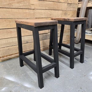 Puede incluir: Dos taburetes de madera negra con una tapa de madera marr&oacute;n. Los taburetes tienen un dise&ntilde;o simple e industrial.