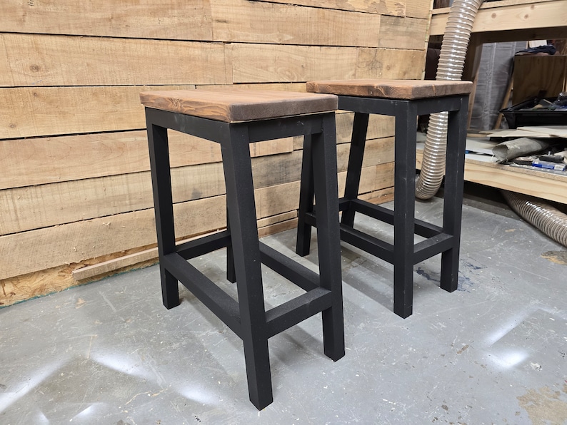 Puede incluir: Dos taburetes de madera negra con una tapa de madera marr&oacute;n. Los taburetes tienen un dise&ntilde;o simple e industrial.