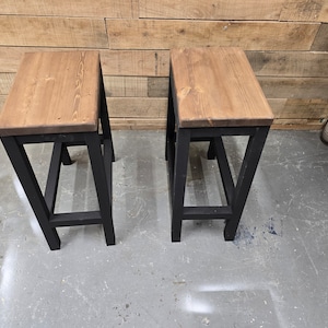 Puede incluir: Dos taburetes de metal negro con tapas de madera. Los taburetes tienen un dise&ntilde;o simple e industrial.