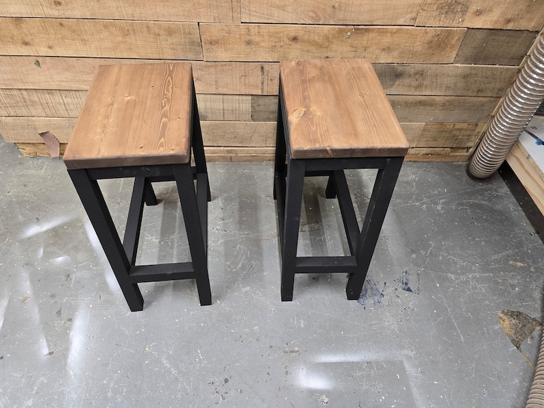 Puede incluir: Dos taburetes de metal negro con tapas de madera. Los taburetes tienen un dise&ntilde;o simple e industrial.