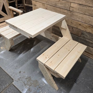 May include: A wooden picnic table with two benches. The table is made of unfinished pine wood and has a simple design. The benches are attached to the table and fold up for easy storage.