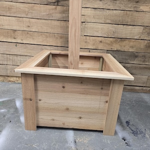 May include: A square wooden planter box with a natural wood finish. The planter has a raised edge and a post extending through the center.