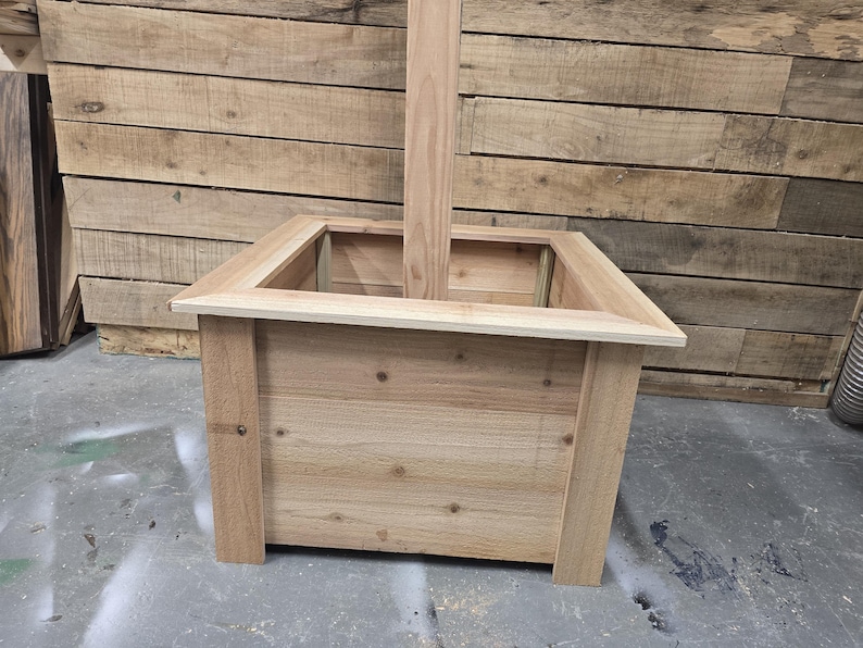 May include: A square wooden planter box with a natural wood finish. The planter has a raised edge and a post extending through the center.