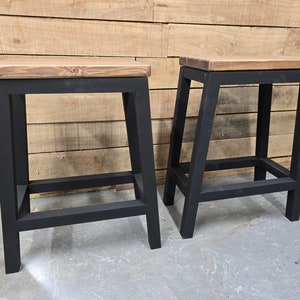 Puede incluir: Dos taburetes de metal negro con tapas de madera. Los taburetes tienen un dise&ntilde;o simple e industrial.