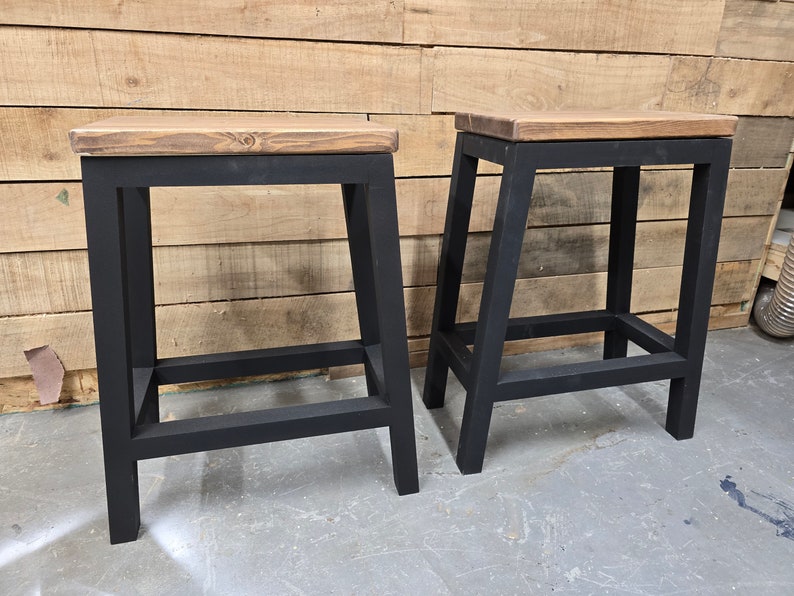 Puede incluir: Dos taburetes de metal negro con tapas de madera. Los taburetes tienen un dise&ntilde;o simple e industrial.