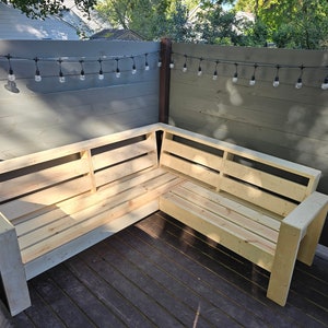 May include: A wooden corner sofa made from unfinished pallet wood. The sofa is on a wooden deck with string lights hanging above.