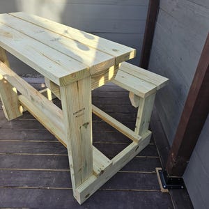 May include: A wooden picnic table with a bench attached to one side. The table is made of unfinished wood and has a natural wood colour. The table is sitting on a wooden deck.