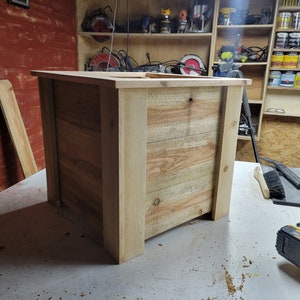 K&ouml;nnte beinhalten: Ein rechteckiger Blumenkasten aus Holz mit naturbelassenem Holzfinish. Der Blumenkasten besteht aus unbehandelten Holzbrettern und hat eine quadratische Form. Der Blumenkasten steht auf einer hellgrauen Oberfl&auml;che.