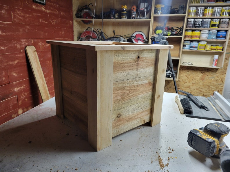 K&ouml;nnte beinhalten: Ein rechteckiger Blumenkasten aus Holz mit naturbelassenem Holzfinish. Der Blumenkasten besteht aus unbehandelten Holzbrettern und hat eine quadratische Form. Der Blumenkasten steht auf einer hellgrauen Oberfl&auml;che.