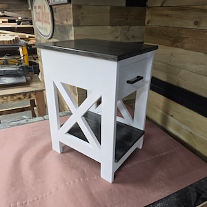 May include: White end table with a black stained top and shelf. The table has a single drawer with a black handle and an X-shaped design on the front.