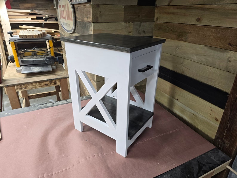May include: White end table with a black stained top and shelf. The table has a single drawer with a black handle and an X-shaped design on the front.