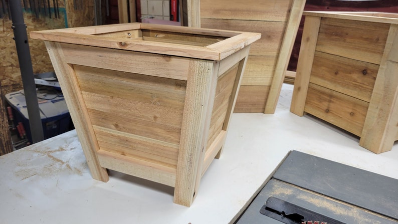 May include: A wooden planter box with a square shape and a light brown finish. The planter has a raised lip around the top and sits on four short legs.