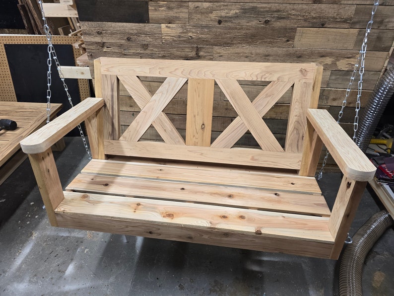 May include: A wooden swing bench with a slatted seat and a decorative X-shaped back. The swing is hanging from two chains.