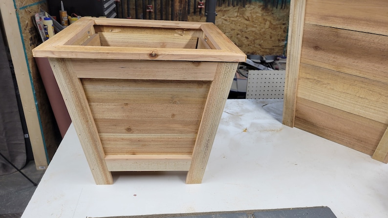 May include: A wooden planter box with a square top and a slightly flared base. The planter is made of natural wood with a light brown color.