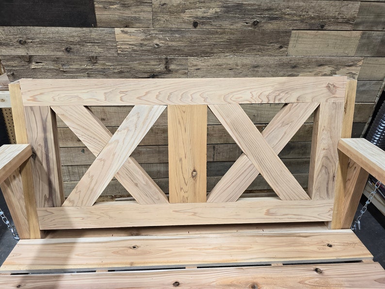 May include: A wooden swing with a slatted seat and a decorative X-shaped back. The swing is made of natural wood and has a rustic look.