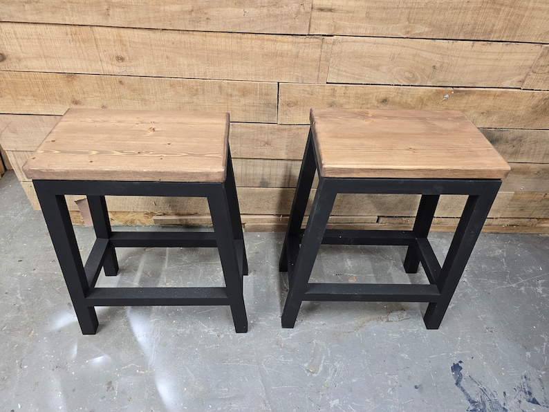 Puede incluir: Dos taburetes de metal negro con tapas de madera. Los taburetes tienen un dise&ntilde;o sencillo con un solo estante.