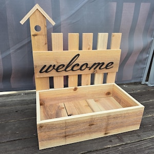 May include: A wooden planter box with a fence-style back and a small birdhouse on top. The word "welcome" is cut out of the wooden sign. The planter is made of light-colored wood and sits on a wooden deck.