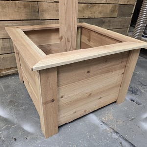 May include: A square wooden planter box with a natural wood finish. The planter has a raised edge and is made of unfinished wood planks.
