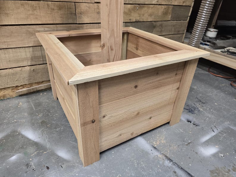 May include: A square wooden planter box with a natural wood finish. The planter has a raised edge and is made of unfinished wood planks.