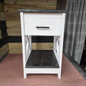 May include: White nightstand with a single drawer and a lower shelf. The drawer has a black handle. The top and shelf are stained dark gray.