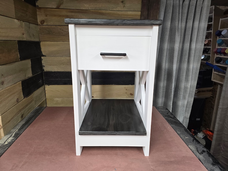 May include: White nightstand with a single drawer and a lower shelf. The drawer has a black handle. The top and shelf are stained dark gray.