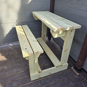 May include: A wooden bench and table set, made of unfinished light wood, with a simple design. The table is attached to a wall and has a bench underneath.