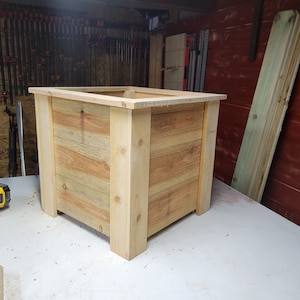 K&ouml;nnte beinhalten: Ein quadratischer Holz-Pflanzkasten aus hellem braunem Holz mit nat&uuml;rlicher Oberfl&auml;che. Der Pflanzkasten steht auf einer wei&szlig;en Oberfl&auml;che.