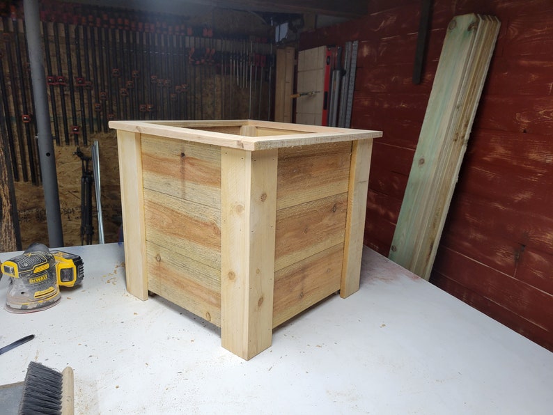 K&ouml;nnte beinhalten: Ein quadratischer Holz-Pflanzkasten aus hellem braunem Holz mit nat&uuml;rlicher Oberfl&auml;che. Der Pflanzkasten steht auf einer wei&szlig;en Oberfl&auml;che.