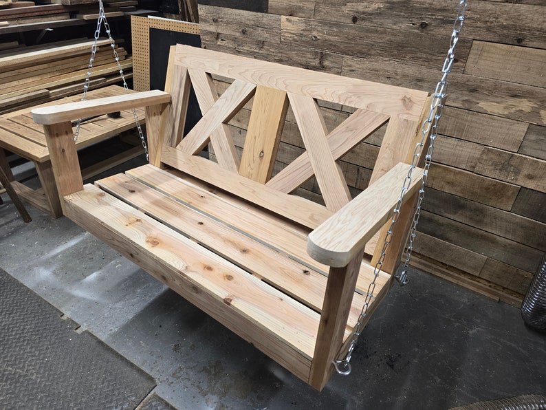 May include: A wooden swing bench with a slatted seat and a decorative X-shaped back. The swing is suspended by two silver chains.