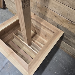 May include: A wooden planter box with a square opening and a vertical wooden post inside. The planter box is made of unfinished wood and has a natural wood color.