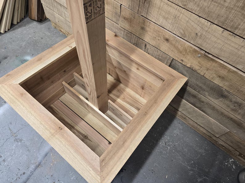 May include: A wooden planter box with a square opening and a vertical wooden post inside. The planter box is made of unfinished wood and has a natural wood color.