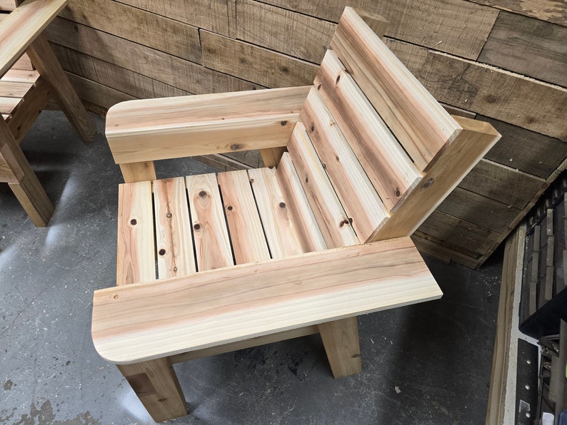 May include: A wooden chair with a slatted back and seat, and a wide armrest on one side. The chair is made of natural wood with a light brown finish.