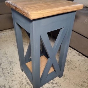 May include: A small, blue, wooden end table with a natural wood top and an X-shaped design on the front. The table is sitting on a gray carpet.